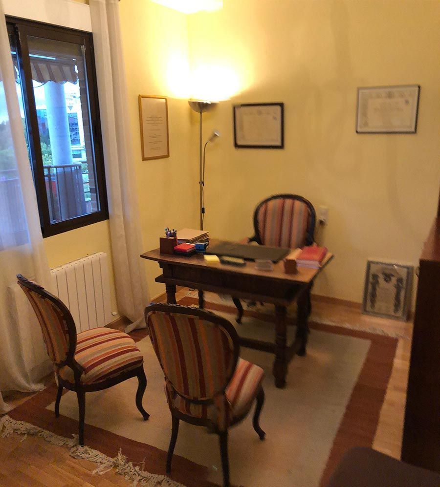 Oficina con escritorio, sillas, ventana, lámpara y documentos enmarcados. Paredes de color amarillo claro, suelo de madera y alfombra.