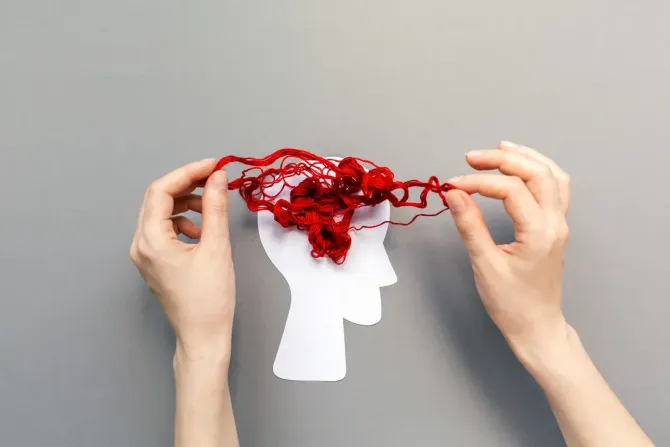 Manos sosteniendo un hilo rojo con forma de cerebro sobre una cabeza de papel blanco sobre un fondo gris.