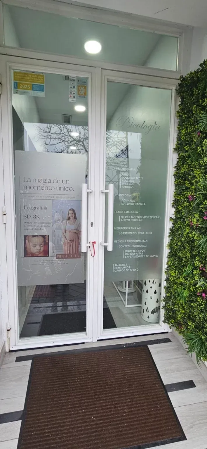 Entrada de cristal con puertas blancas, carteles y un felpudo marrón bajo un toldo verde.