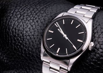 Un reloj plateado con esfera negra reposa sobre una muñeca de cuero negro.