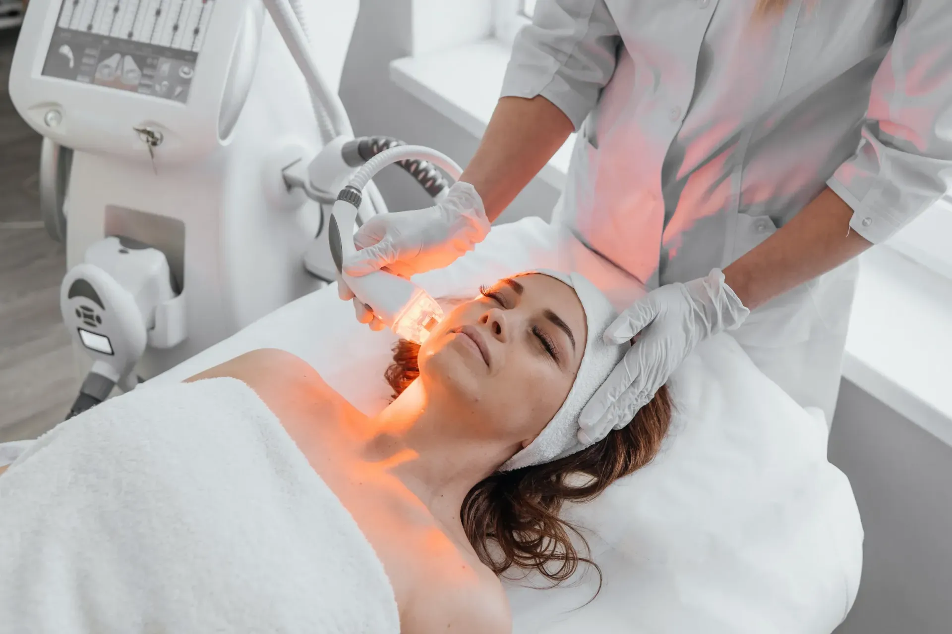 Mujer recibiendo un tratamiento facial con terapia de luz roja en un spa. Un técnico le sostiene el dispositivo sobre el rostro mientras está tumbada en una camilla blanca.