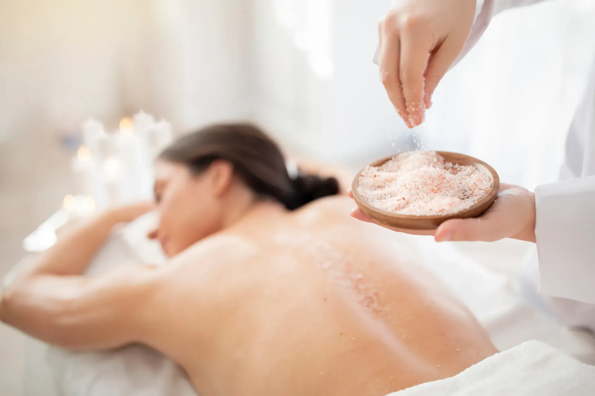 Una persona recibe un exfoliante con sal en la espalda en un spa; una mano rocía sal rosada desde un recipiente de madera.