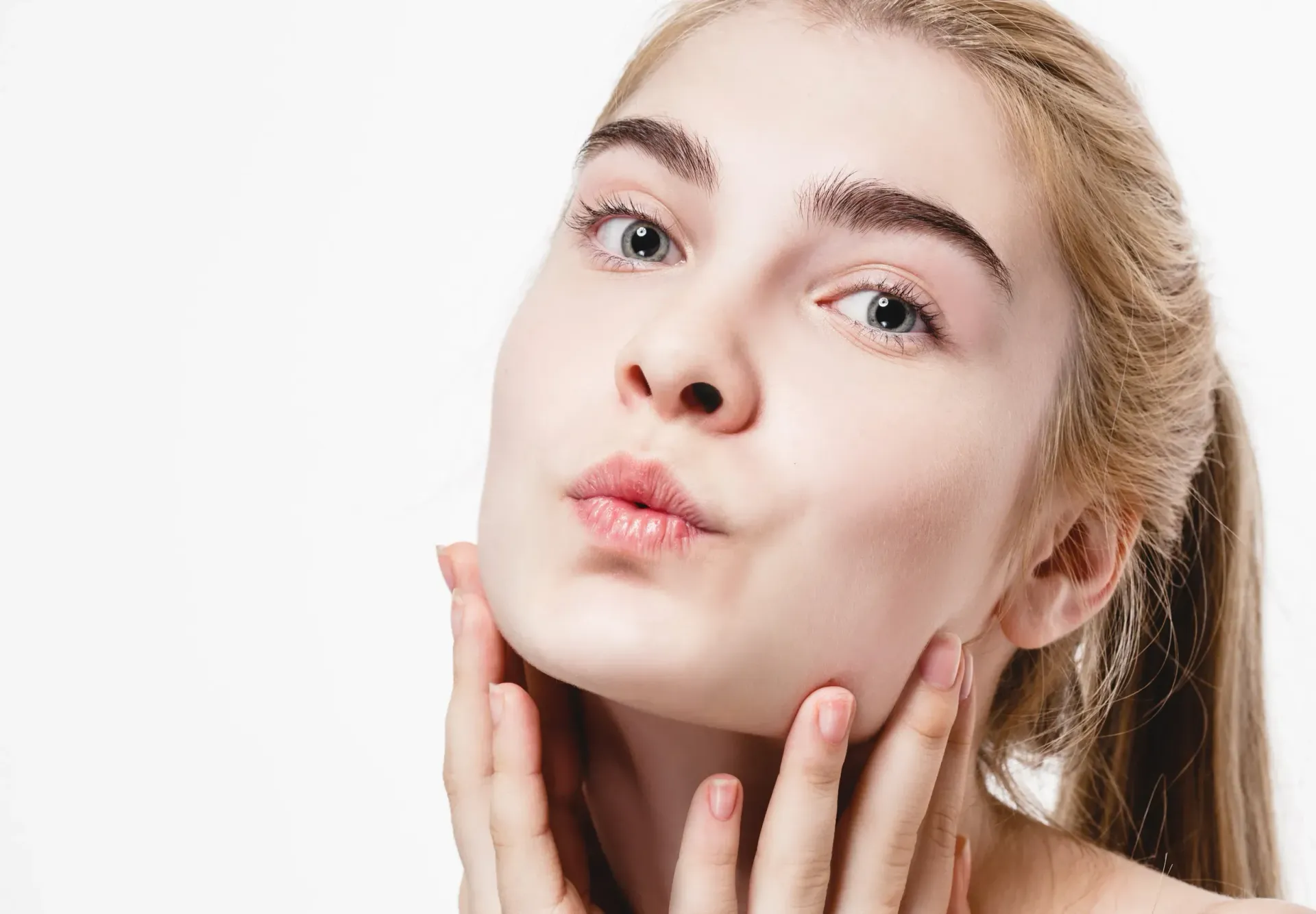 Mujer rubia de piel clara frunciendo los labios, tocándose las mejillas con ambas manos, fondo blanco.