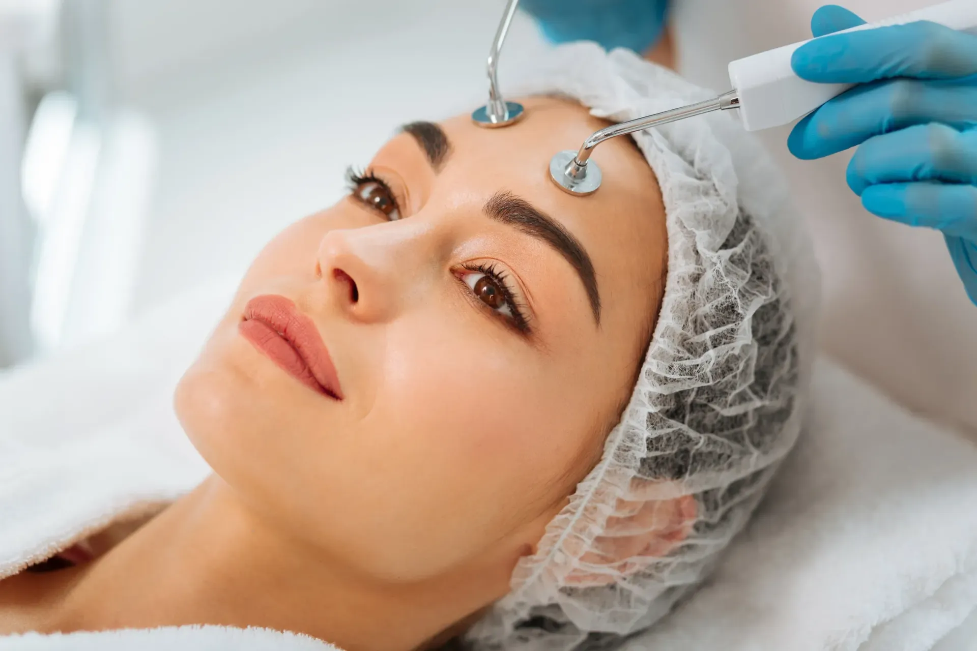 Una mujer recibe un tratamiento facial con electrodos en la frente en un spa. Lleva una redecilla para el pelo y mira hacia arriba.