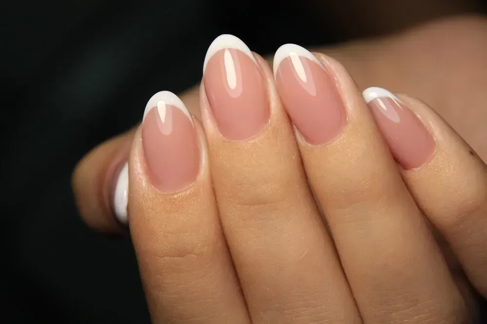 Mano con uñas de forma ovalada que presenta una manicura francesa clásica: base nude con puntas blancas.