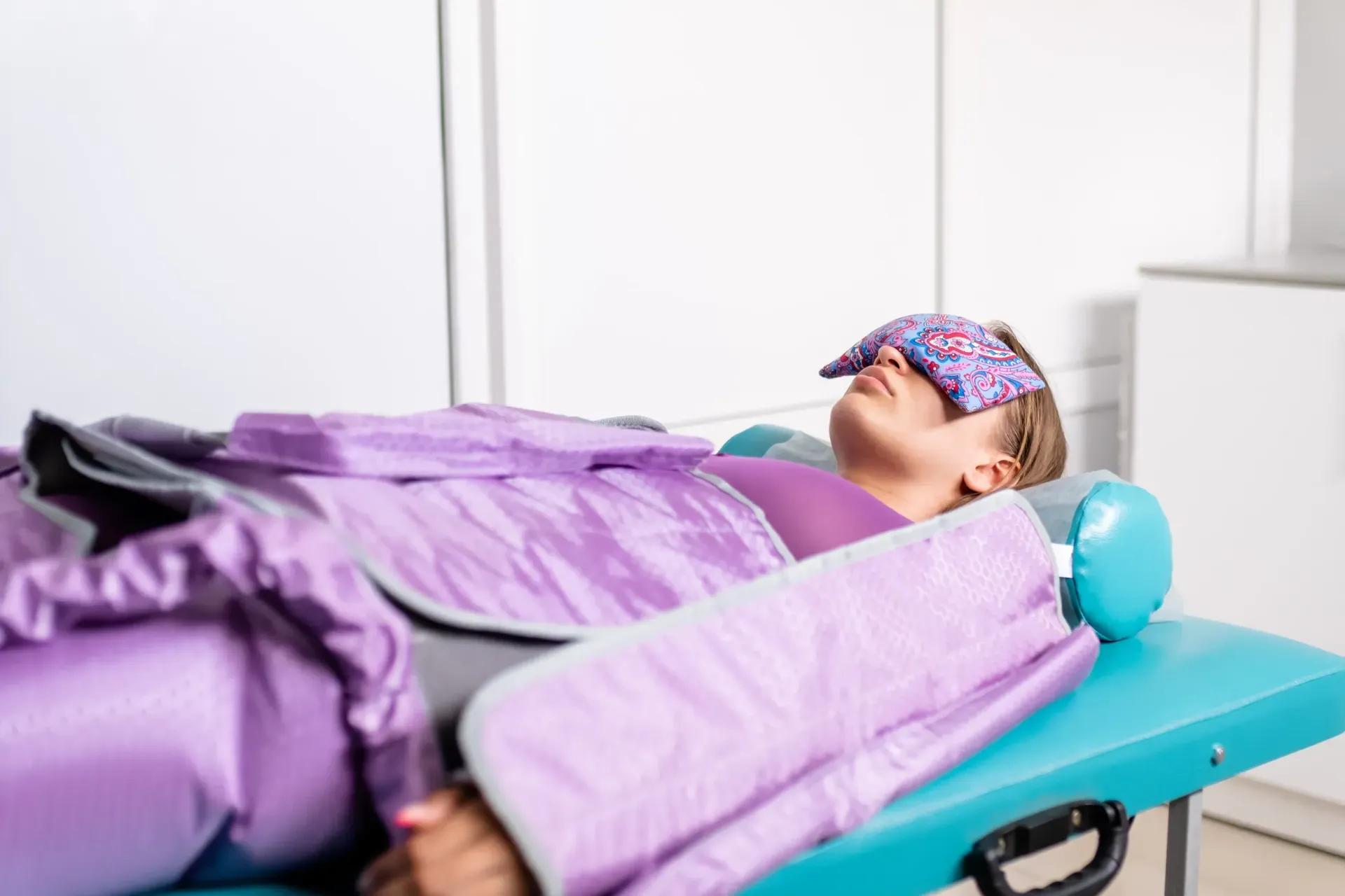 Mujer acostada en una cama médica turquesa, sometiéndose a un tratamiento de drenaje linfático con prendas de compresión moradas y una máscara para los ojos.