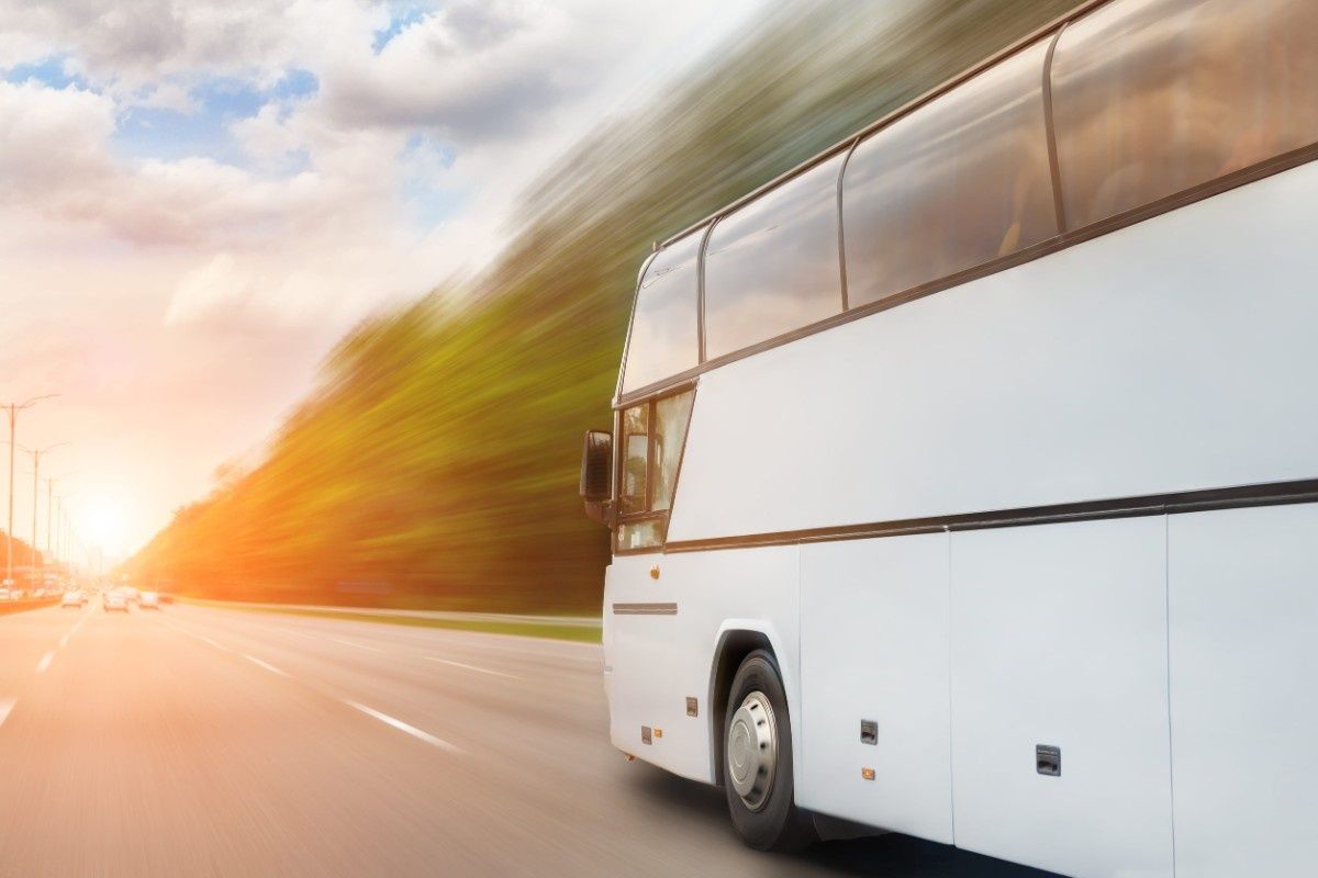 Un autobús blanco circula por una autopista al atardecer, con un fondo de árboles y luces borrosos por el movimiento.