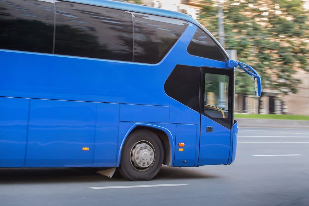 Un autobús azul circula por una carretera asfaltada con un fondo borroso de árboles y edificios.