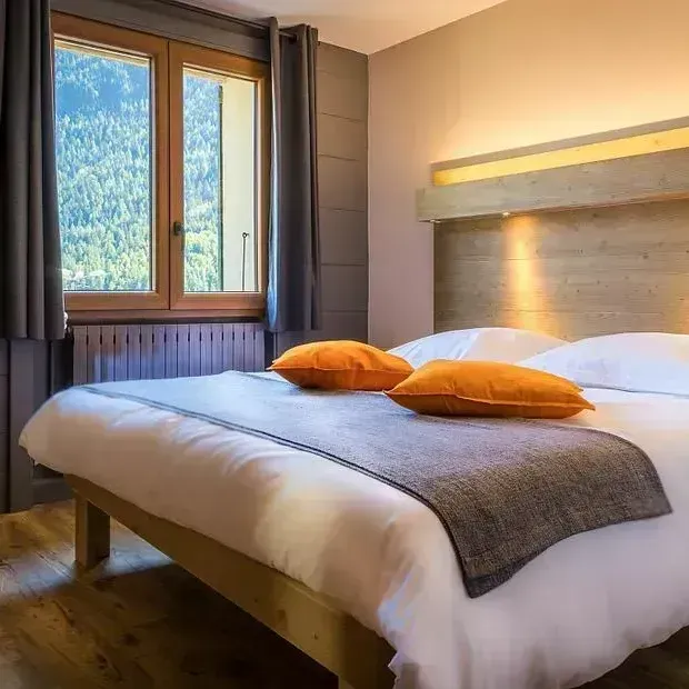 Chambre avec un lit double, une couverture grise, des oreillers orange et une fenêtre avec vue sur la montagne.