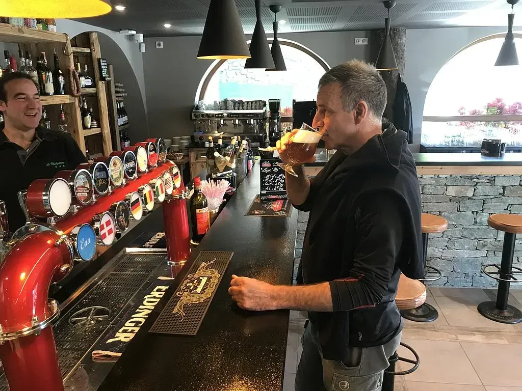 Un homme boit une bière dans un bar. Un barman est derrière le comptoir. Intérieur sombre avec des tireuses et des bouteilles.