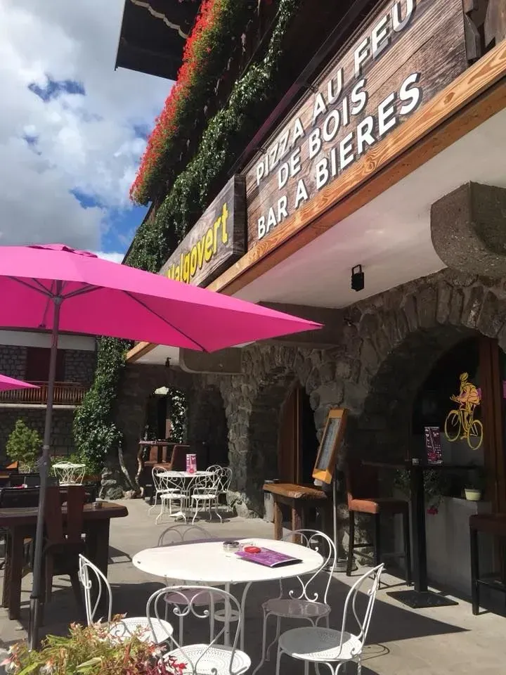 Façade du restaurant avec parasols roses, tables blanches et arches en pierre. Enseigne :  Pizza au feu de bois, bar à bières.
