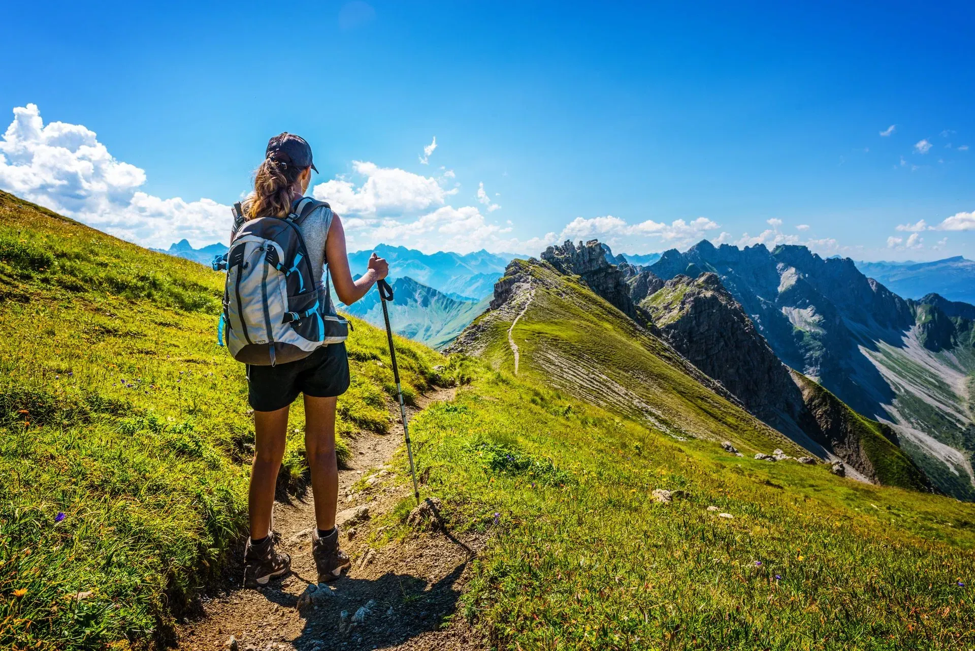 Femme randonnant sur une crête montagneuse verdoyante, sac au dos et utilisant des bâtons de marche, sous un ciel bleu.