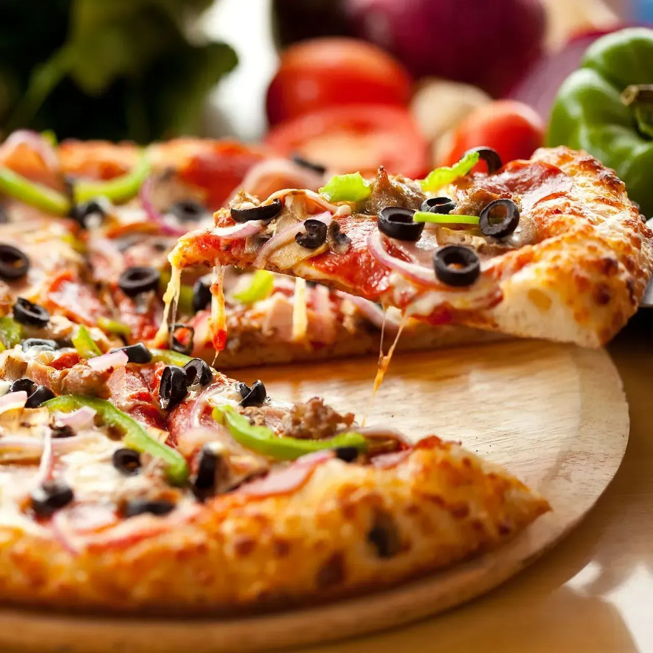 Pizza dont une part est soulevée, garnie d'olives, de poivrons et d'oignons, sur une planche en bois.