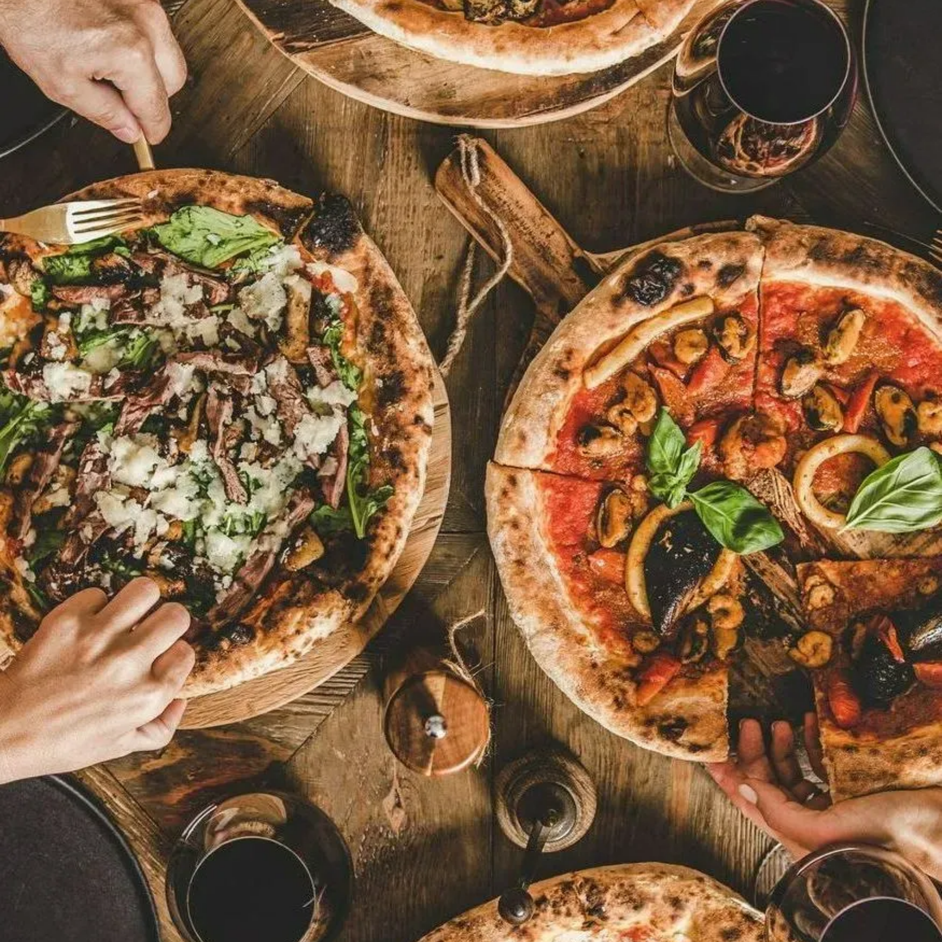 Vue aérienne d'une table en bois avec plusieurs pizzas, des mains qui se servent en parts et des boissons.
