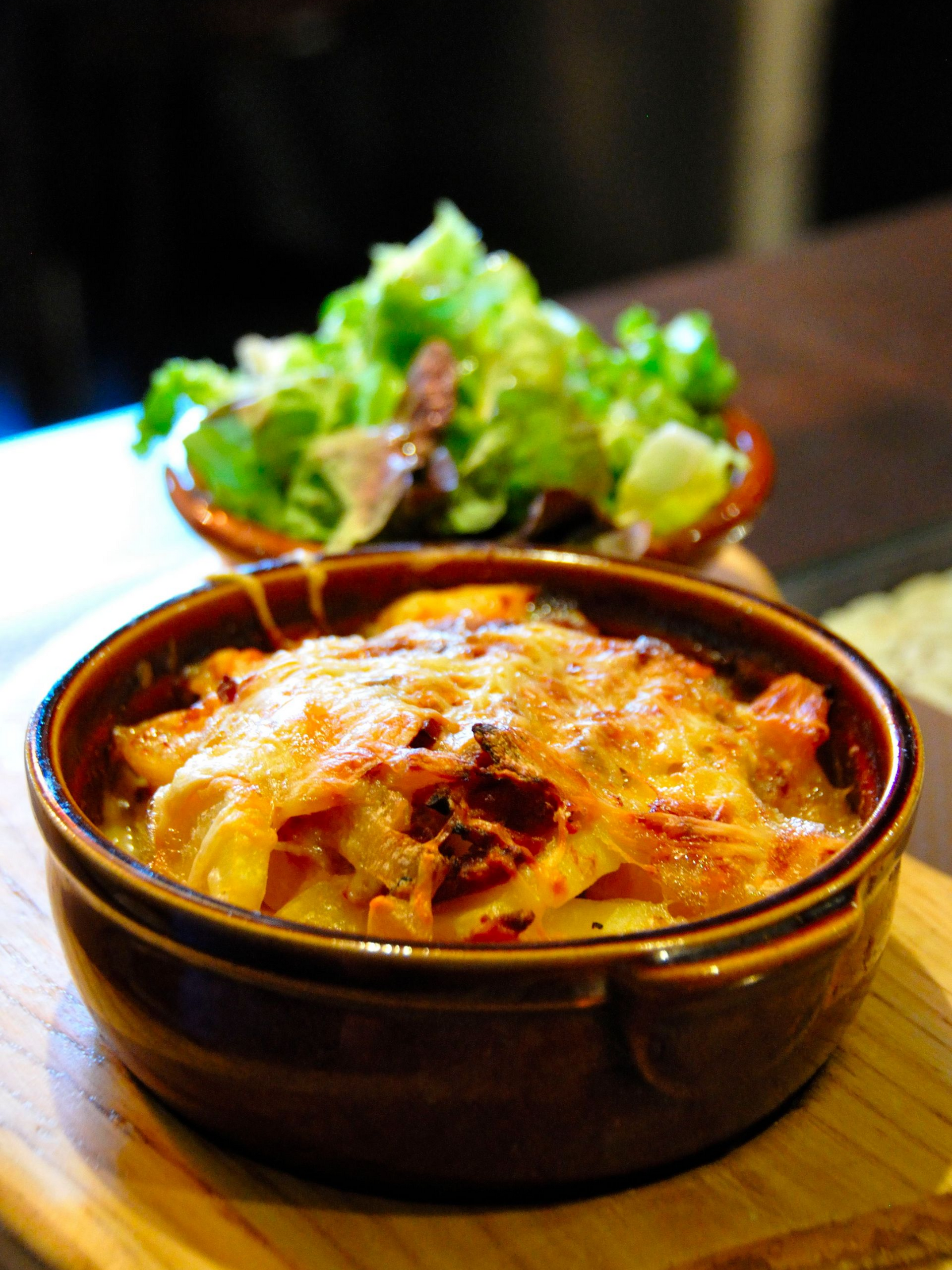 Gratin de pommes de terre au fromage fondu, servi dans un plat brun avec une salade d'accompagnement sur une planche en bois.