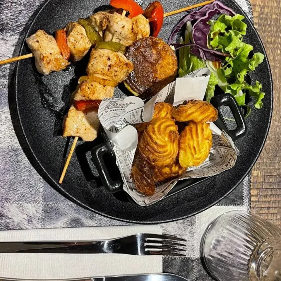 Brochette de poulet avec légumes, pommes de terre frites, pâte feuilletée et salade, servie sur une assiette noire.