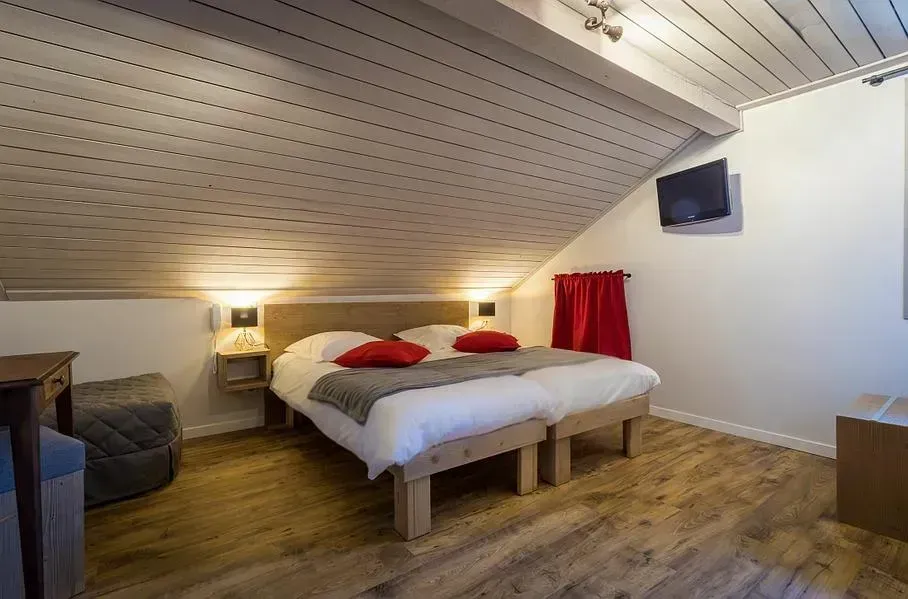 Chambre avec deux lits jumeaux, parquet et plafond mansardé. Touches de rouge, petite télévision et lampes de chevet.