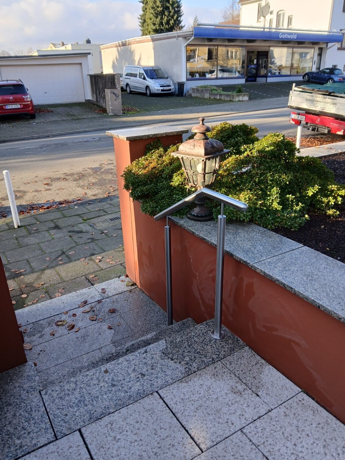 Ein metallenes Geländer führt über Steinstufen neben einer roten Mauer und einer dekorativen Außenleuchte an einem Gebäude entlang.