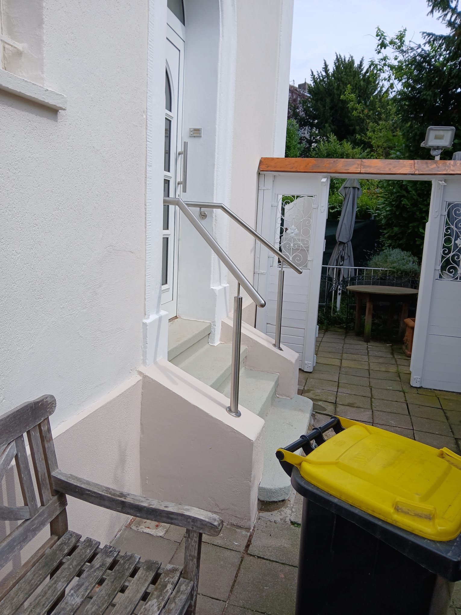 Ein beigefarbener Gebäudeeingang mit drei Betonstufen, einem Metallgeländer, einer Holzbank und einem gelben Mülleimer auf einer Terrasse.