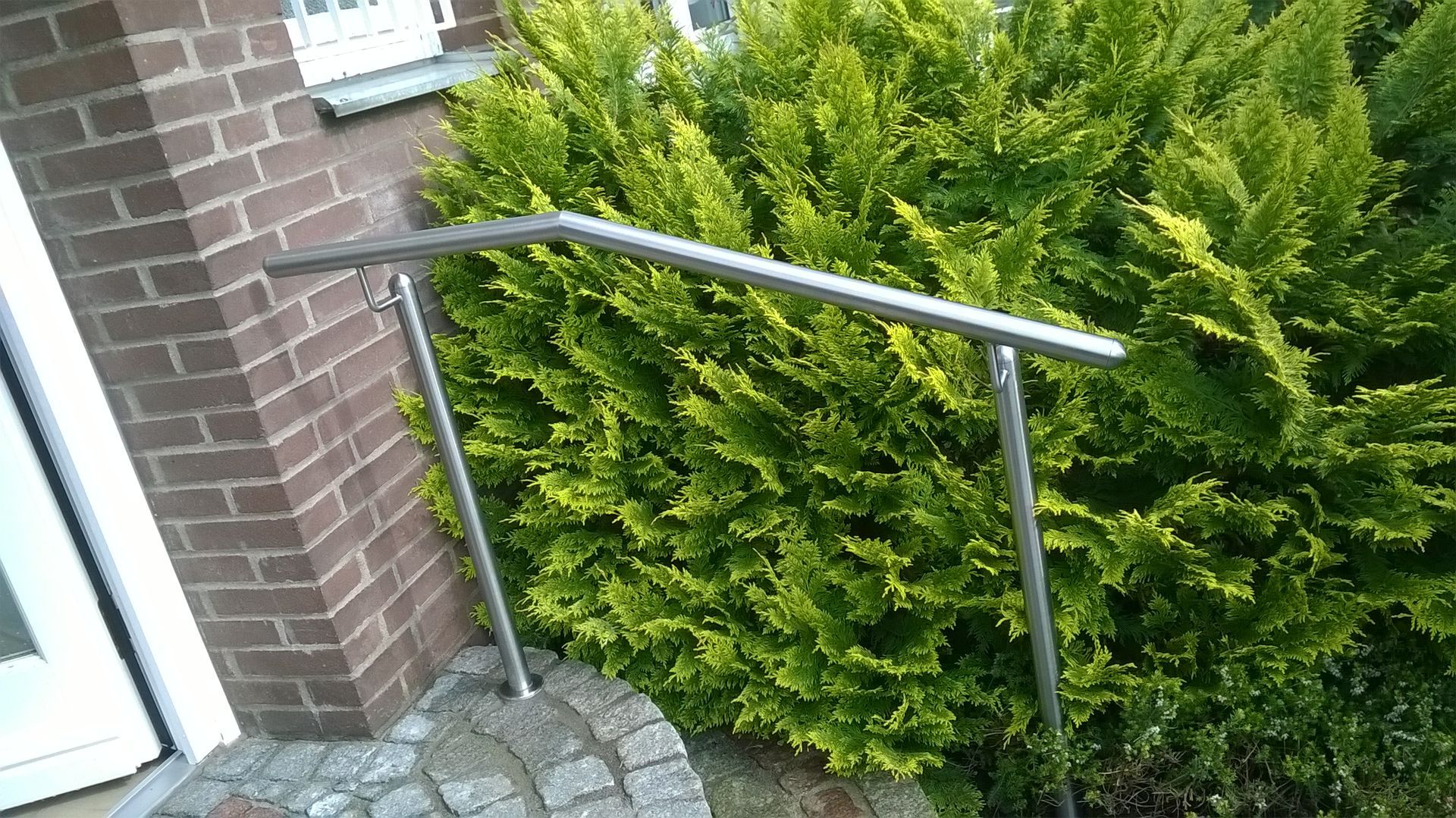 Ein schlichtes, leicht geneigtes Metallgeländer, das im Freien neben einem Backsteingebäude und einem leuchtend grünen Busch angebracht ist.