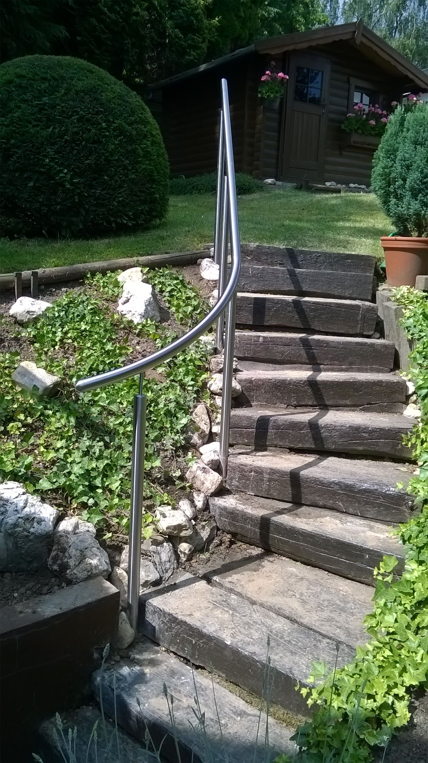 Eine schmale Treppe aus Betonsteinen in einem Garten, mit einem einzelnen Edelstahlgeländer, die zu einem Holzschuppen führt.