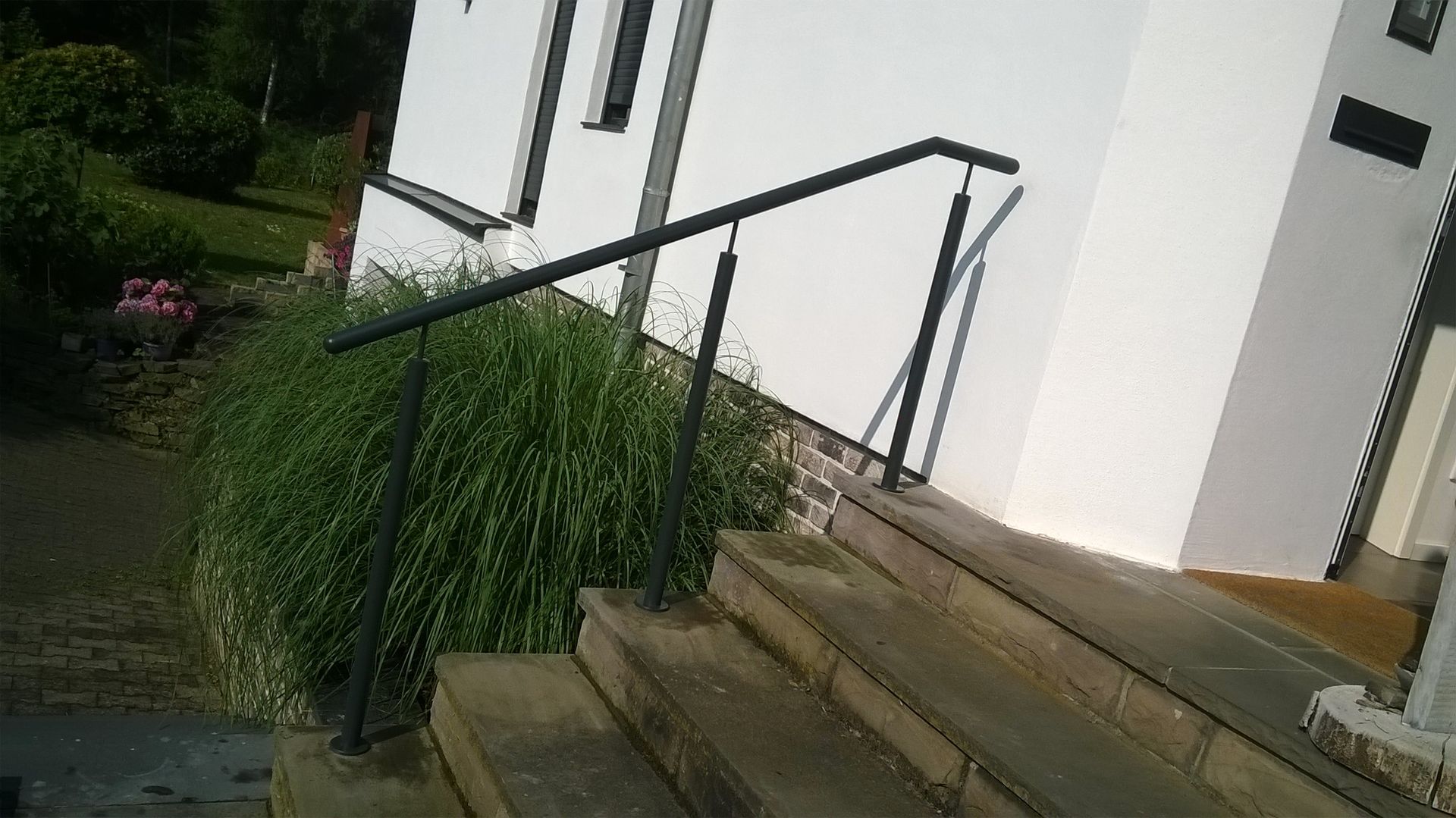 Ein dunkles Metallgeländer ist entlang einer steinernen Außentreppe angebracht, die zu einem weißen Gebäudeeingang führt.