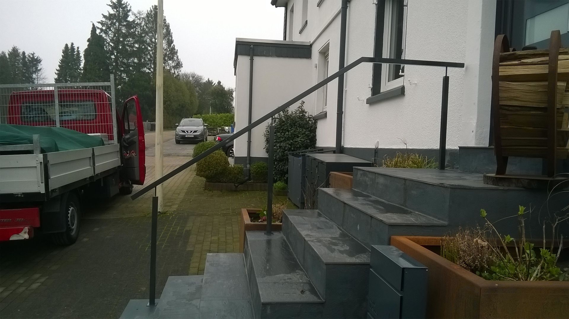 Eine graue Steintreppe führt zum Hauseingang, daneben befindet sich ein einfacher Metallhandlauf und ein in der Nähe geparkter Lastwagen.