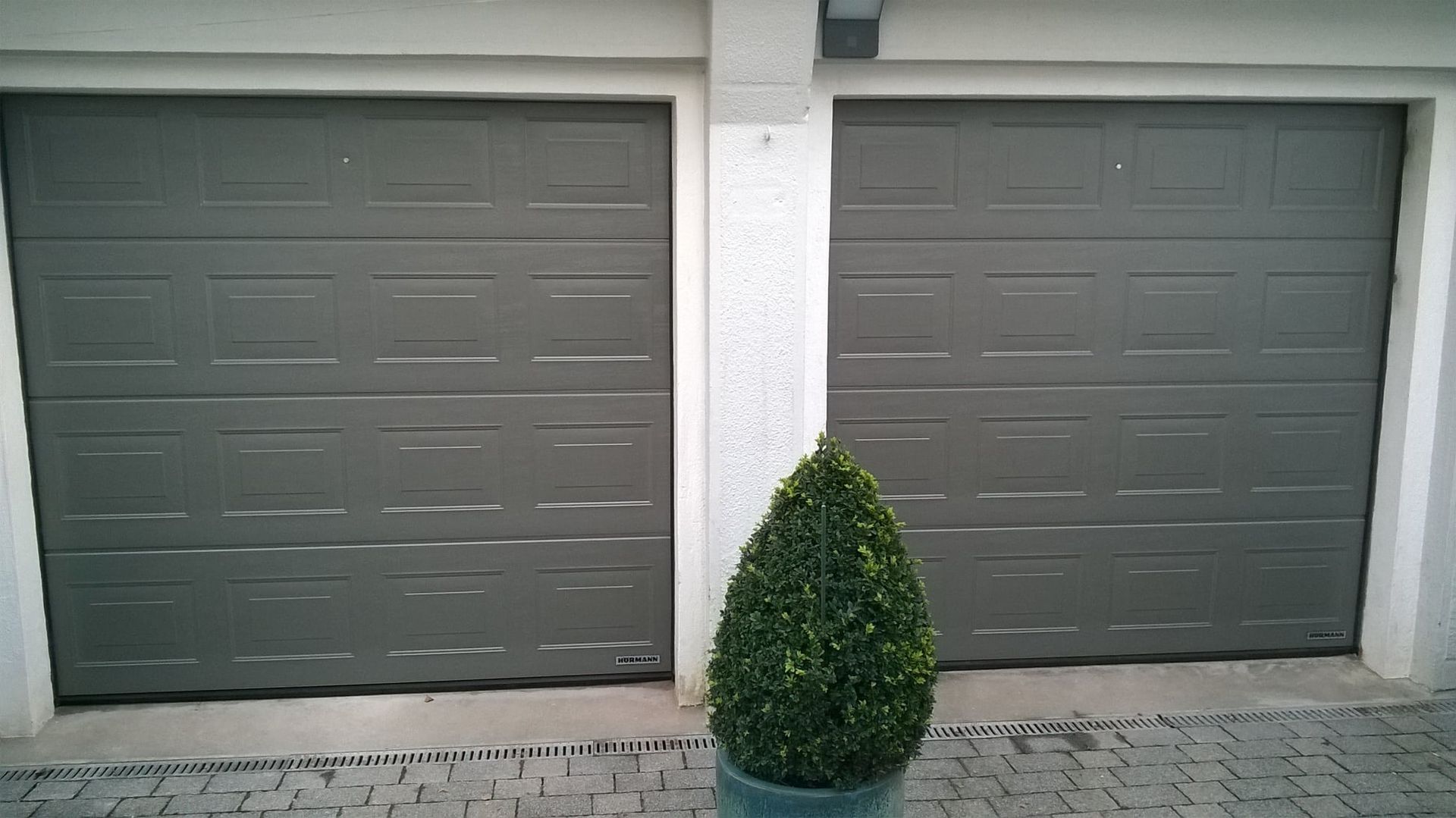 Zwei graue, holzgetäfelte Garagentore nebeneinander, getrennt durch eine weiße Säule, im Vordergrund ein kleiner, kegelförmiger Strauch.