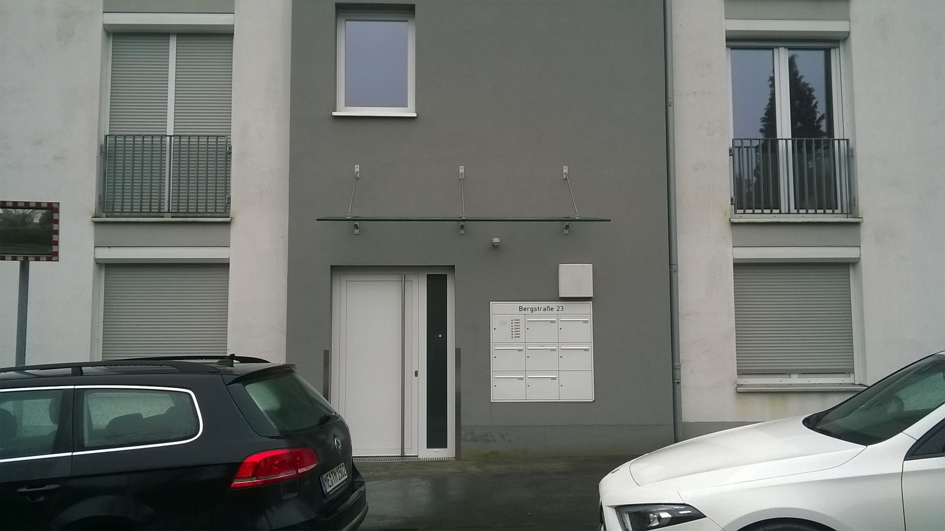 Der Eingang eines modernen Mehrfamilienhauses mit weißer Tür, grauer Fassade, Briefkästen und geparkten Autos im Vordergrund.