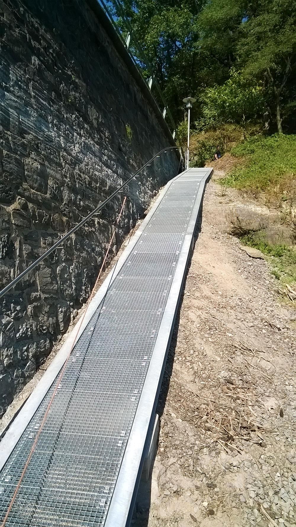Ein Metallweg, der neben einer Steinmauer einen Hügel hinaufführt.
