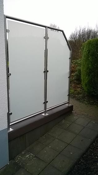 Auf einem Gehweg neben einem Gebäude steht ein Edelstahlzaun mit Milchglas.