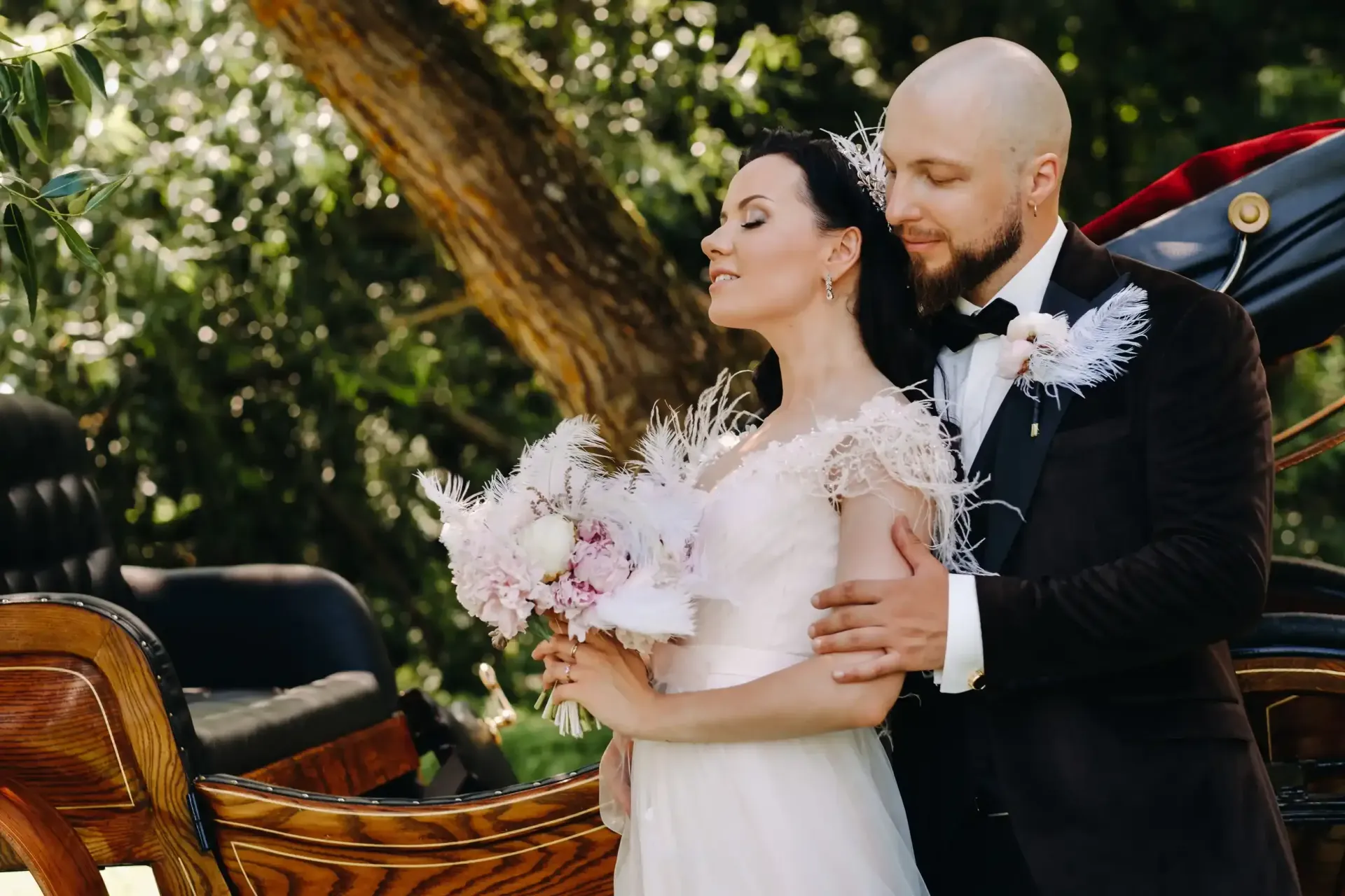 Los novios se abrazan junto a un carruaje, ella sostiene flores, él viste un traje oscuro, entorno al aire libre.