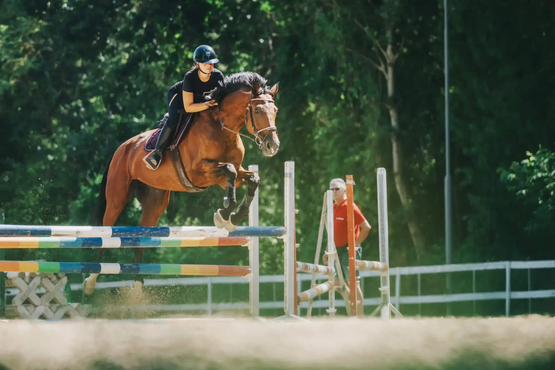 Un jinete sobre un caballo marrón salta una valla colorida en una pista al aire libre. Una persona observa.