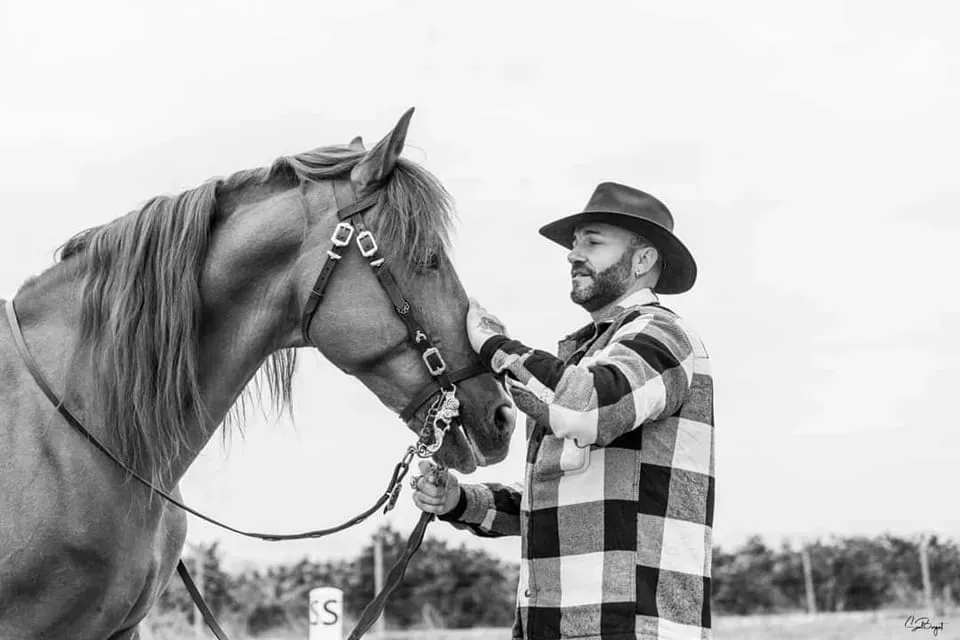 Un hombre con sombrero de vaquero ajusta las bridas de su caballo; ambos están al aire libre. Foto en blanco y negro.