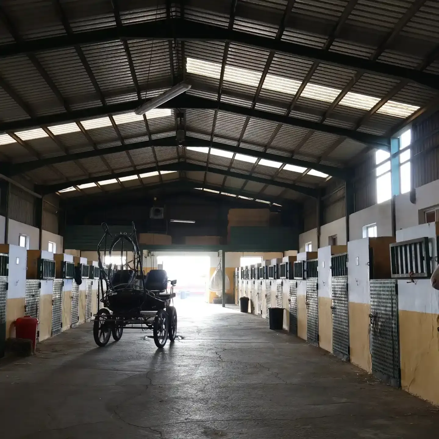 Dentro de un establo con boxes para caballos. Un carruaje está en el centro y la luz del sol brilla desde el fondo.
