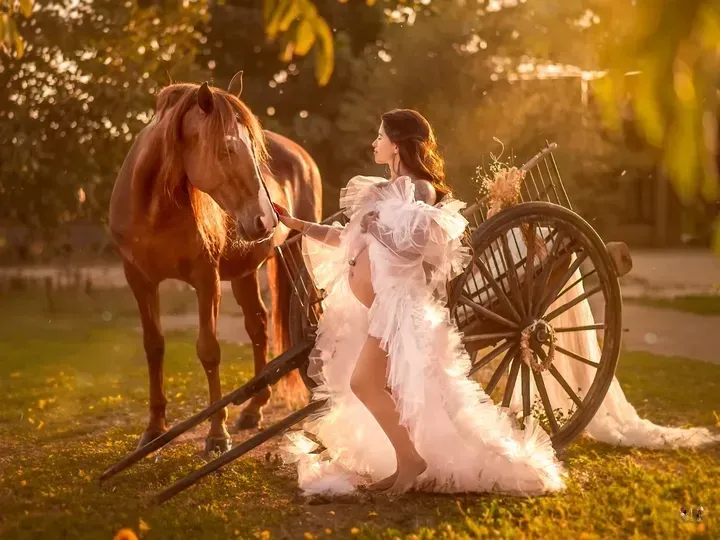 Mujer embarazada con vestido blanco, cerca de un caballo marrón y un carro de madera, al aire libre bajo la luz del sol.