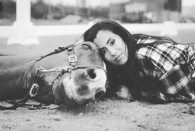Mujer en un caballo marrón, inclinando la cabeza sobre un camino de tierra, con un sombrero negro, en el campo.