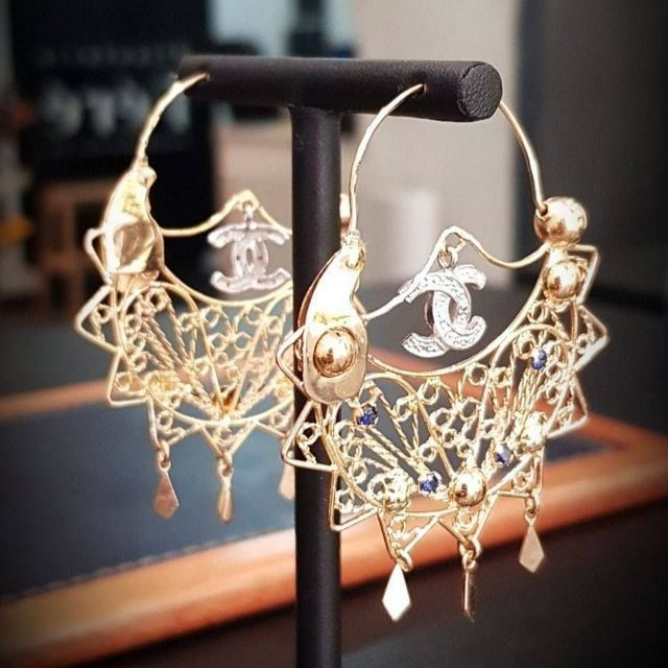 Boucles d'oreilles créoles en or, ornées de filigranes et du logo Chanel, présentées sur un support noir.