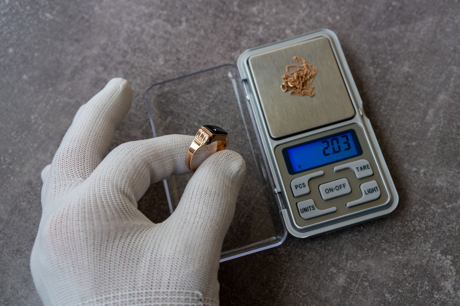 Main gantée tenant une bague en or près d'une balance numérique affichant 20,3, avec de petits objets en or sur la balance.