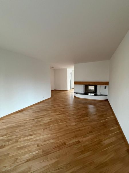 Ein leeres Wohnzimmer mit hellen Holzböden, weissen Wänden und einem modernen Kamin mit hölzernem Kaminsims.