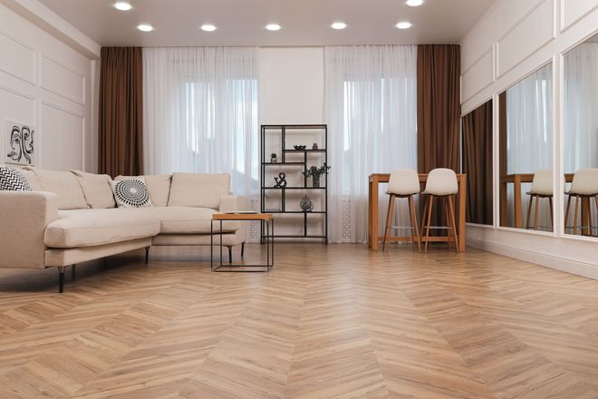 Ein modernes Wohnzimmer mit Fischgrätparkett, einem beigefarbenen Ecksofa, einem schwarzen Regal und einem hohen Bartisch.
