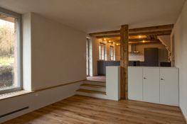 Innenansicht eines Zimmers mit Holzboden, einer kleinen Treppe, einem hölzernen Stützpfeiler und einem erhöhten Küchenbereich.