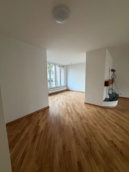 Ein leerer Raum mit hellen Holzböden, weissen Wänden, grossen Fenstern und einem eingebauten Kamin.