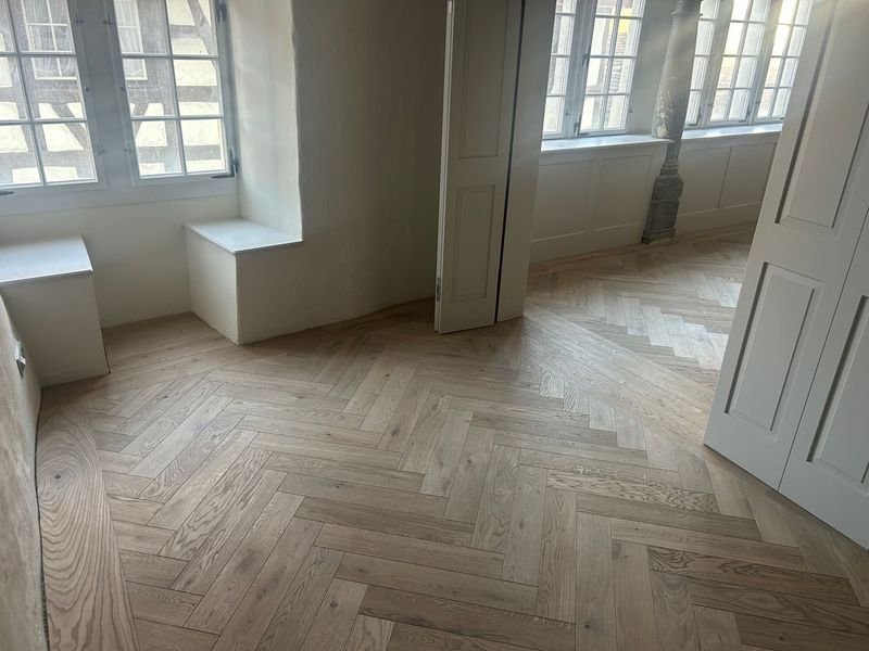 Ein Zimmer mit hellen Fischgrätparkettböden, weissen Wänden, eingebauten Fenstersitzen und mehreren Fenstern.