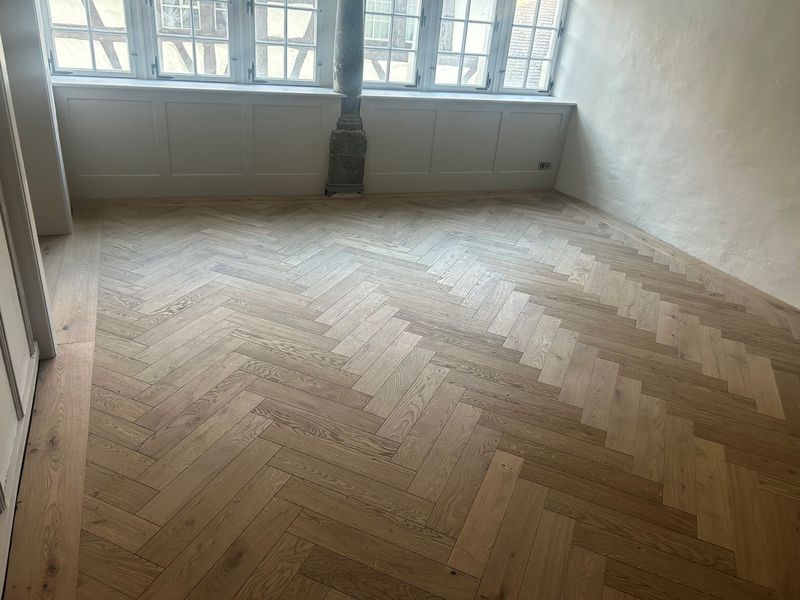 Ein Zimmer mit hellen, im Fischgrätmuster verlegten Holzböden, weissen Wänden und grossen Fenstern mit einem Holzofen.