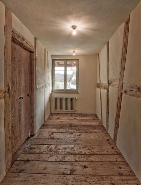 Ein rustikaler Flur mit freiliegenden Holzbalken, naturbelassenen Putzwänden, breiten Holzdielen und einem Fenster am Ende.