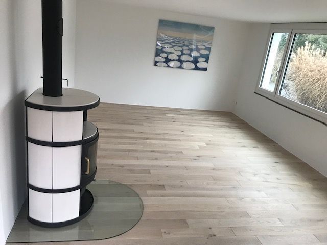 In einem minimalistisch eingerichteten Raum mit hellen Holzböden steht ein moderner, weiss-schwarzer Holzofen auf einer Glasplatte.