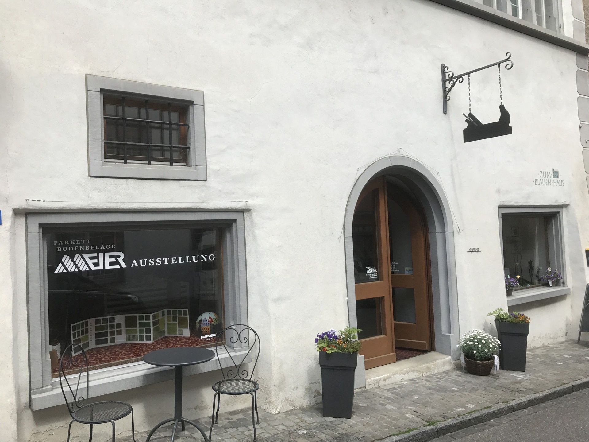 Eine weisse Gebäudefassade mit einem Schaufenster mit der Aufschrift „MEIER AUSSTELLUNG“, einem zentralen Bogeneingang und schmiedeeisernen Verzierungen.