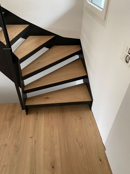 Eine moderne Treppe mit Holzstufen und dunklen Metallstützen vor einer weißen Wand neben hellem Holzboden.