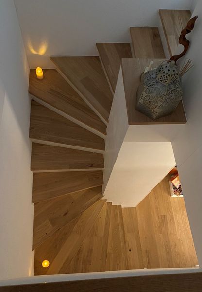 Eine Draufsicht auf eine helle Holztreppe, die sich in einem weiss getünchten Treppenhaus mit leuchtenden, runden Wandleuchten nach unten windet.